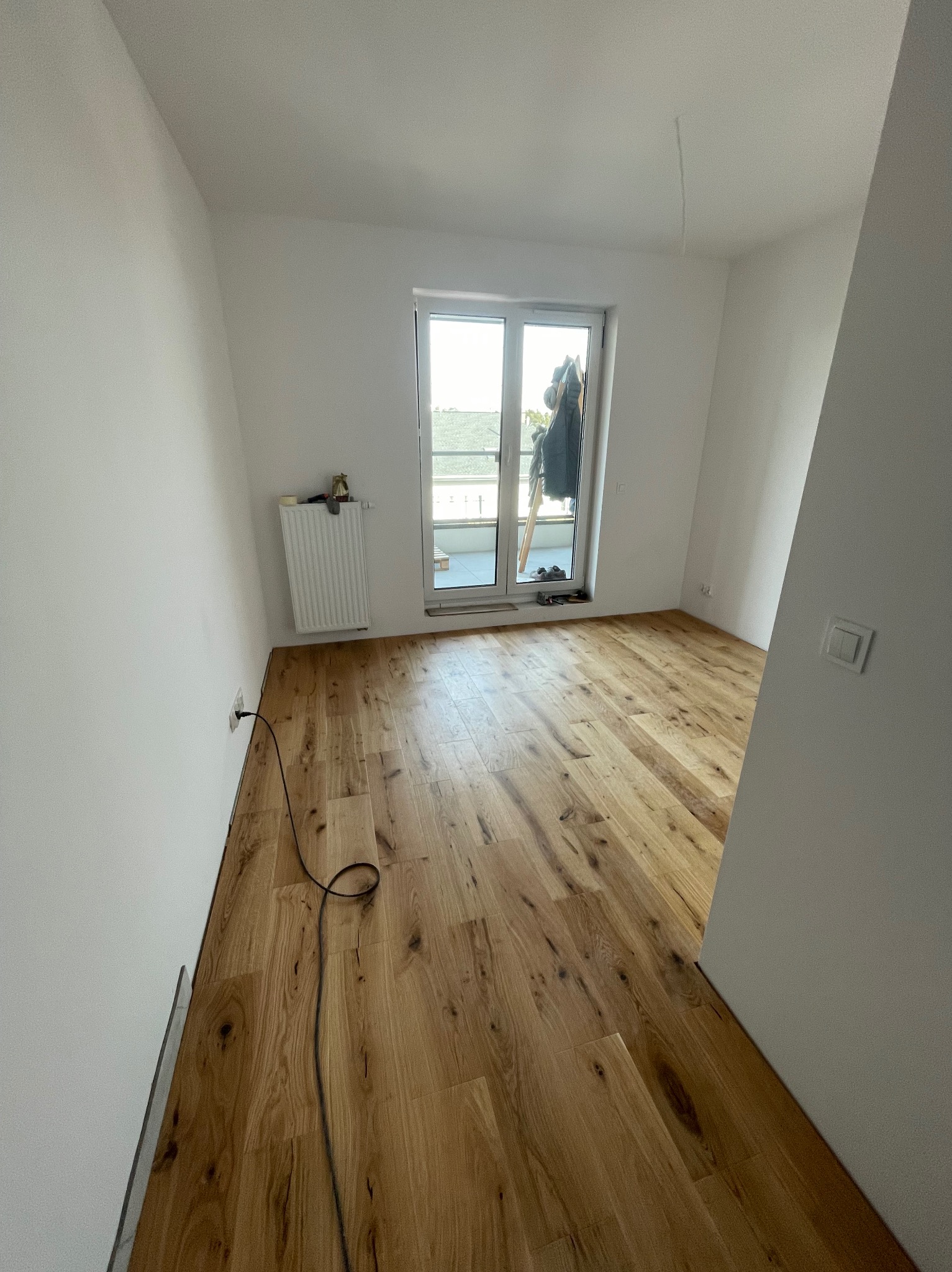 Jasne, drewniane panele podłogowe w nowym pokoju z białymi ścianami i grzejnikiem. Widok na balkon przez otwarte drzwi balkonowe. Kabel elektryczny na podłodze.