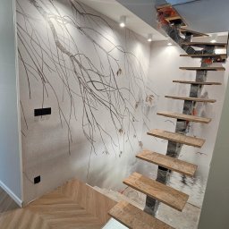 EXACT HOME FIRMA REMONTOWO BUDOWLANA DAWID LISOWSKI - Nowoczesne schody z drewnianymi stopniami i metalową konstrukcją, częściowo zabezpieczoną folią, na tle ściany z motywem drzewa i parkietu ułożonego w jodełkę.