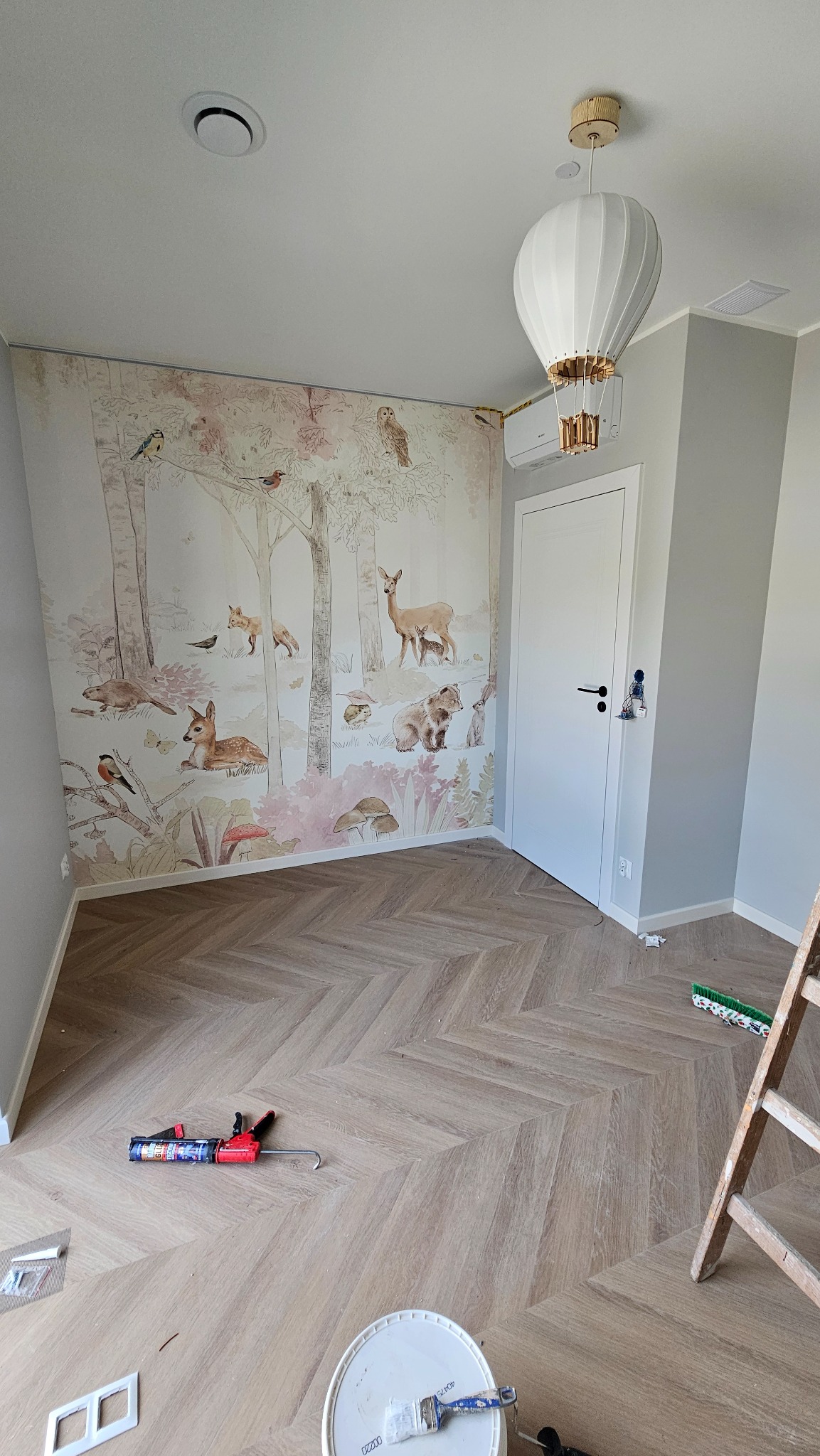 Pokój dziecięcy po remoncie: wzorzysta tapeta z motywem leśnym, parkiet ułożony w jodełkę, dekoracyjna lampa w kształcie balonu. Widoczne narzędzia malarskie.