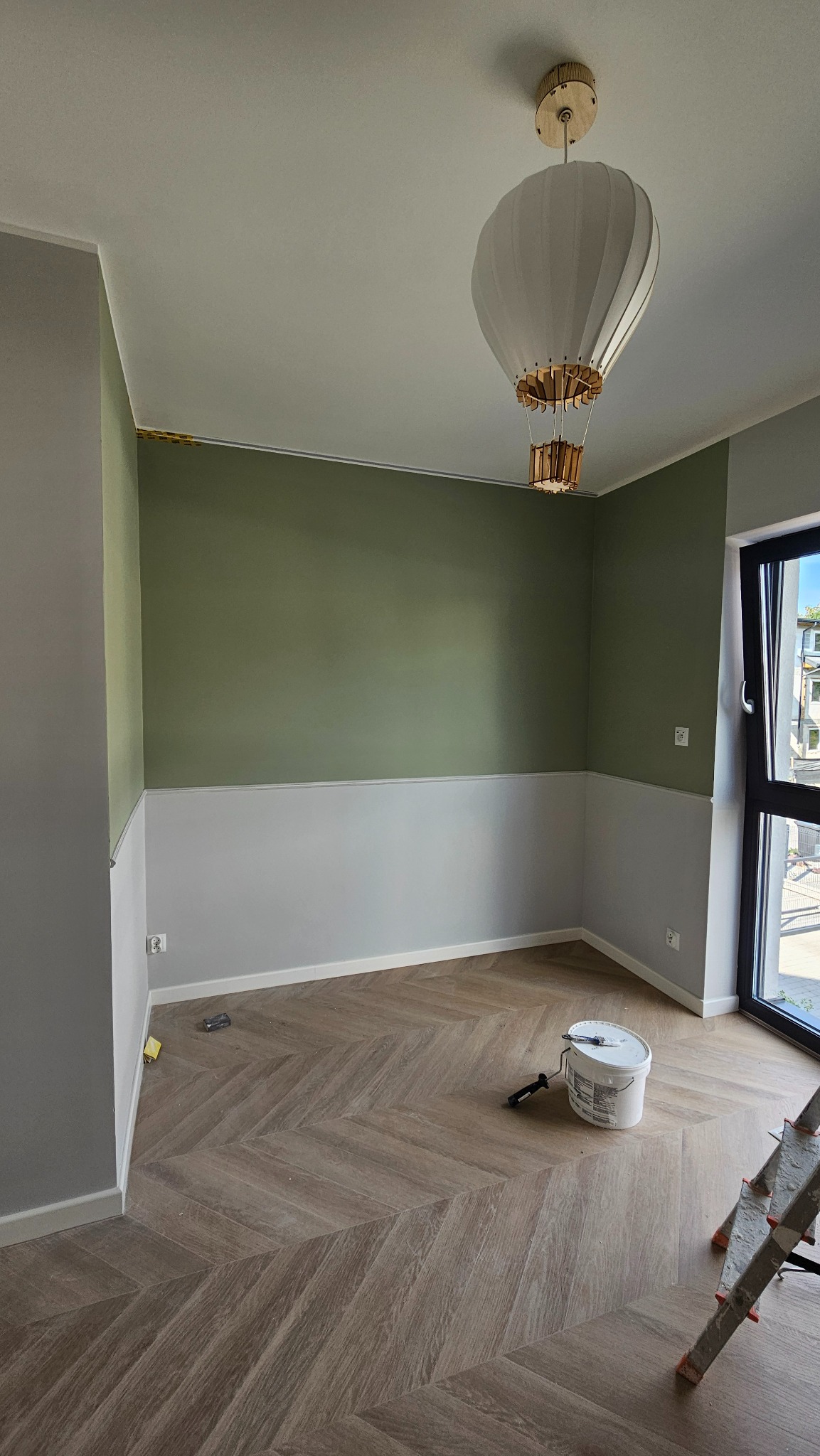 Pokój po malowaniu z zieloną górną częścią ścian i białą dolną, parkiet w jodełkę, drabina malarska i wiadro z farbą na podłodze, lampa w kształcie balonu.