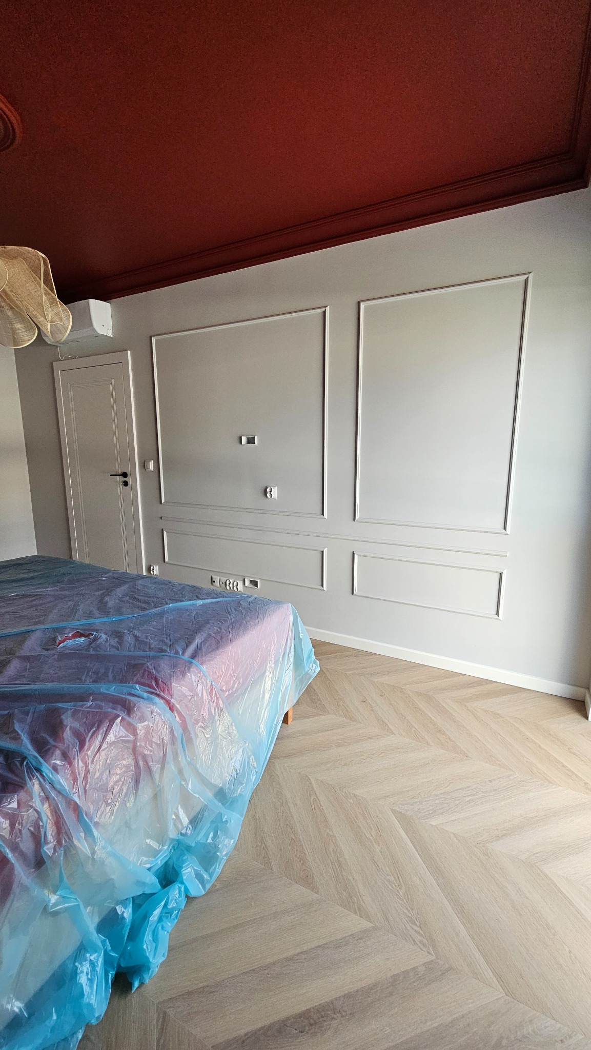 Pokój po remoncie: bordowy sufit, szare ściany z dekoracyjnymi listwami, parkiet w jodełkę. Łóżko zabezpieczone folią malarską.