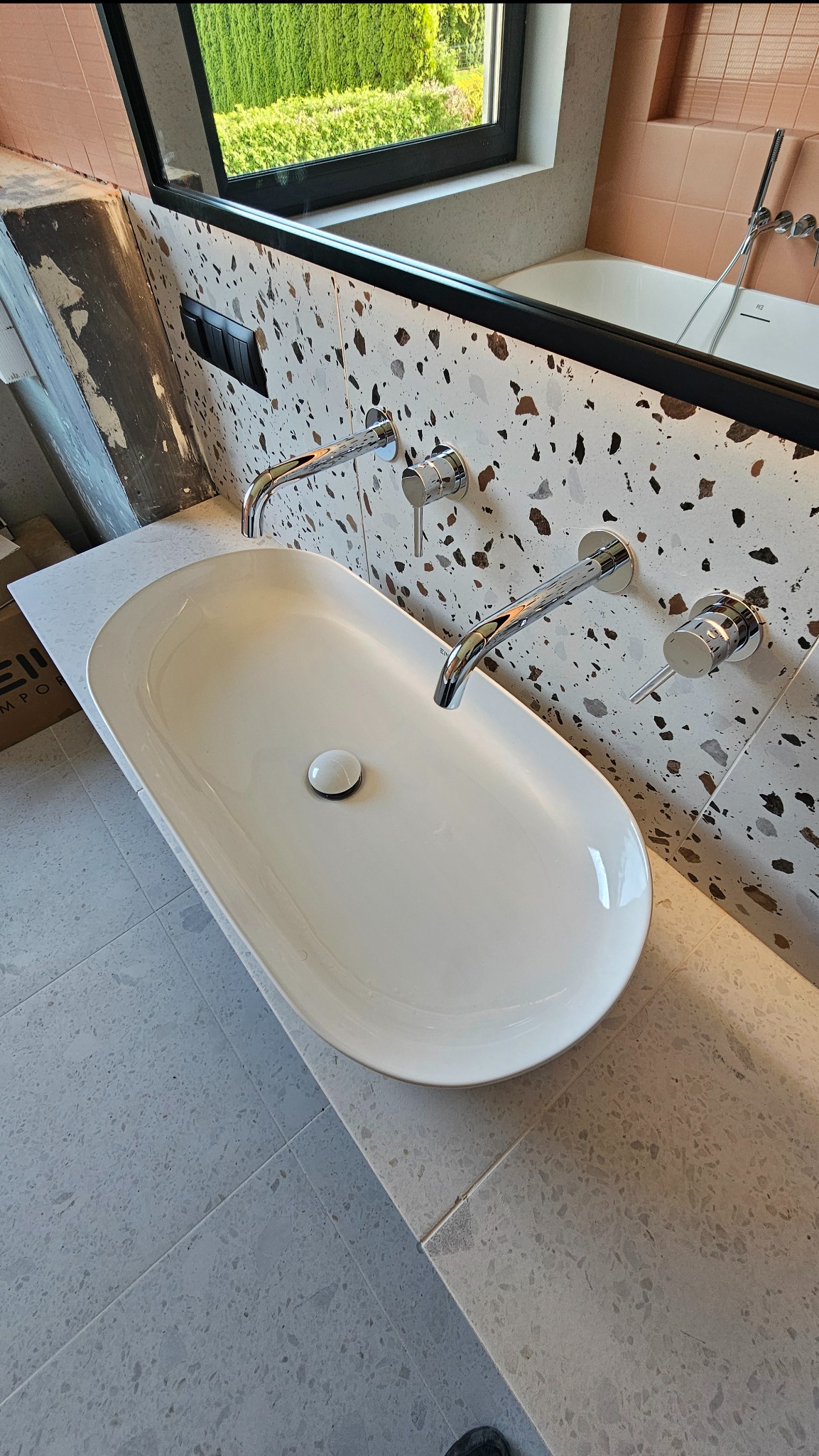 Biała, owalna umywalka nablatowa z chromowaną armaturą na tle ściany wykończonej płytkami lastryko. Widoczna wanna i okno w tle. Nowoczesna aranżacja łazienki.