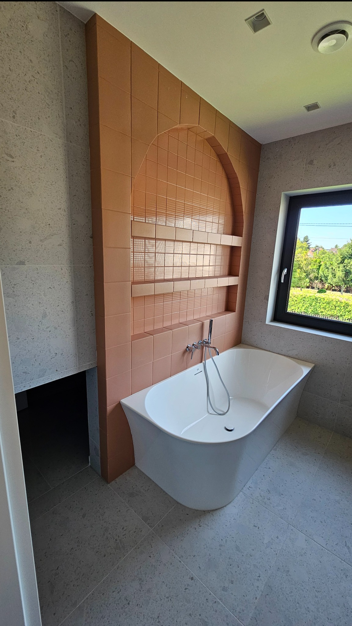 Nowoczesna łazienka z wanną i ścianą z terakotowych płytek z wnękami. Szare płytki podłogowe i okno z widokiem na zieleń. Minimalistyczny design.