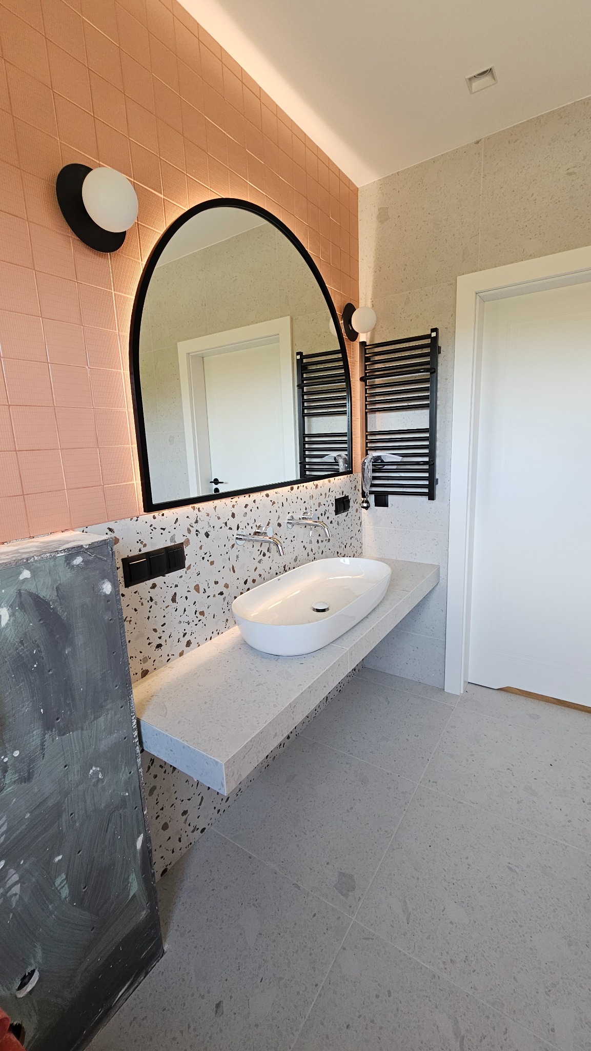 Nowoczesna łazienka z umywalką nablatową, lustrem w czarnej ramie i grzejnikiem drabinkowym. Ściany wyłożone płytkami w odcieniach różu i beżu.