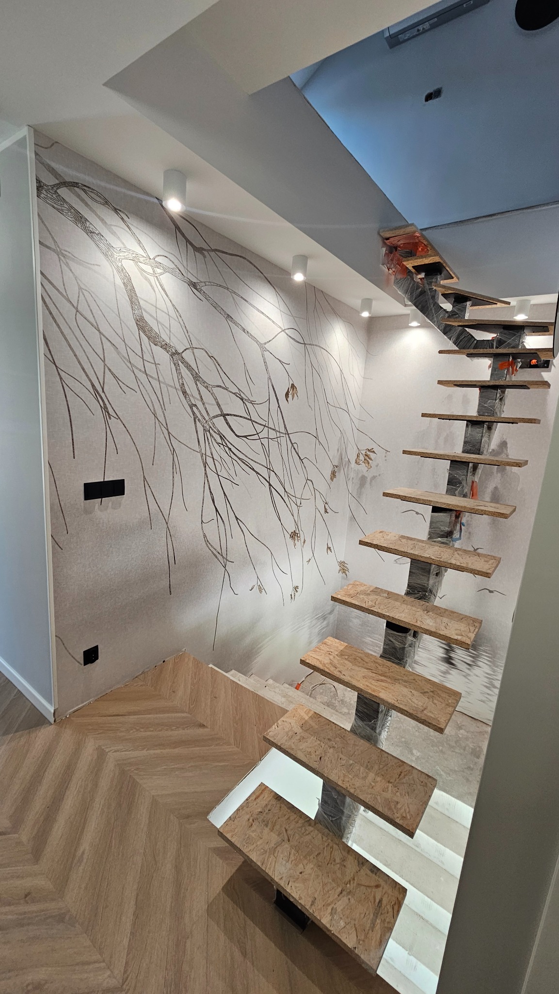 Nowoczesne schody z drewnianymi stopniami i metalową konstrukcją, częściowo zabezpieczoną folią, na tle ściany z motywem drzewa i parkietu ułożonego w jodełkę.