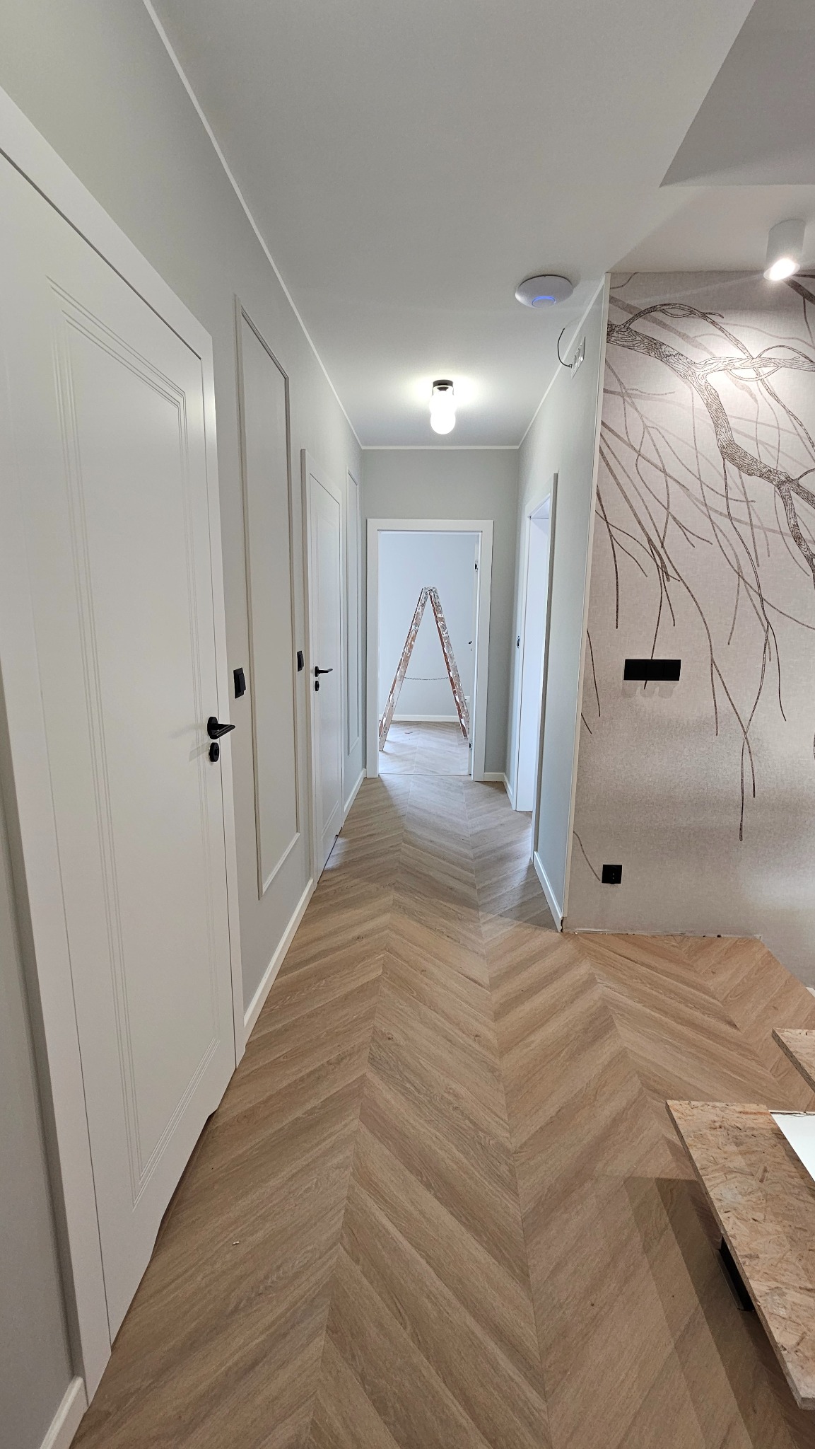 Korytarz po remoncie: parkiet w jodełkę, białe drzwi z czarnymi klamkami, ściana z motywem drzewa. W tle drabina malarska, sugerująca trwające prace wykończeniowe.