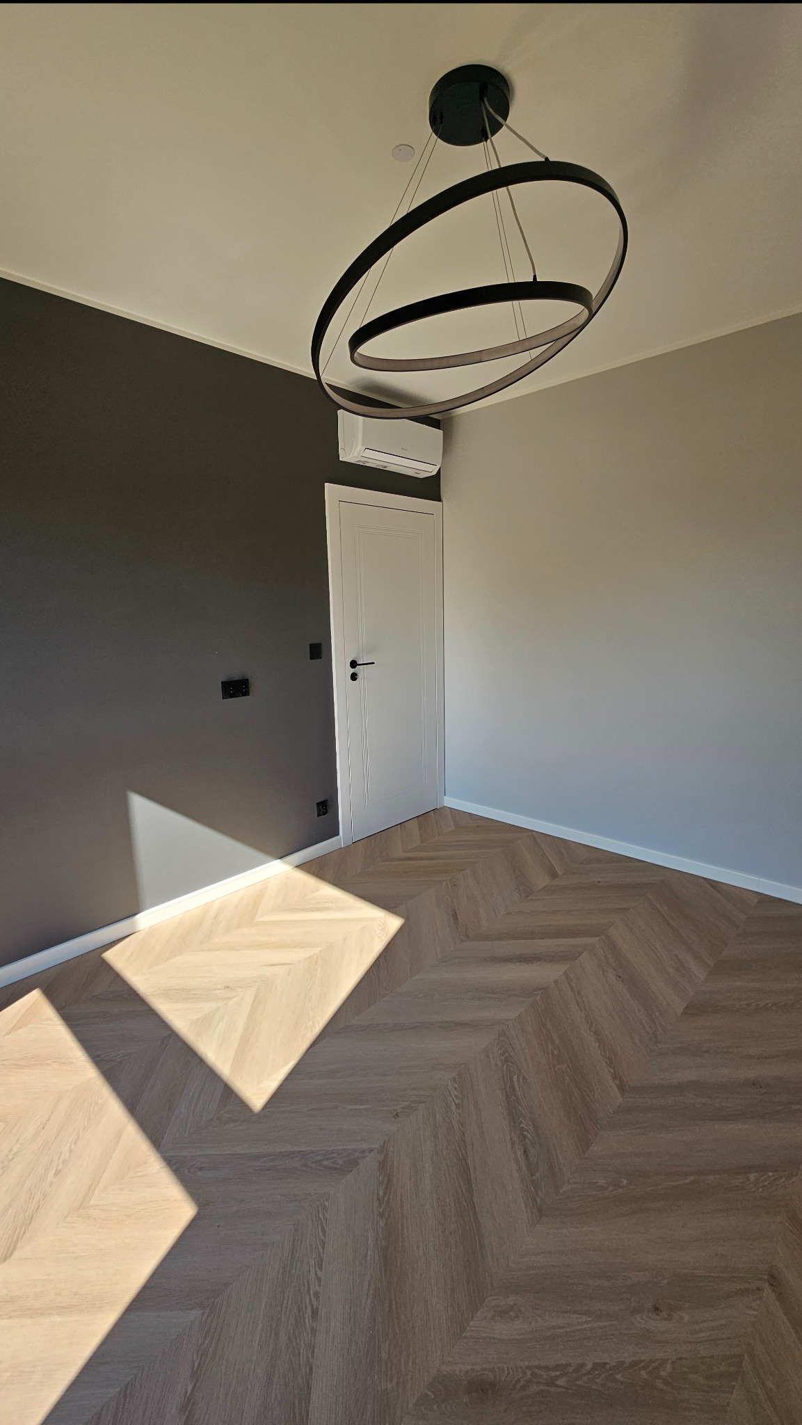 Jasny parkiet ułożony we wzór jodełki francuskiej w pokoju z szarą ścianą i nowoczesną lampą. Widoczny klimatyzator nad białymi drzwiami.