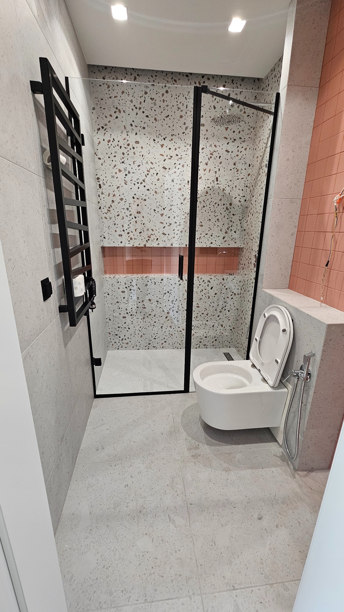 Nowoczesna łazienka z kabiną prysznicową typu walk-in z czarnymi ramami, wiszącą toaletą i grzejnikiem drabinkowym. Płytki w stylu lastryko i akcenty w kolorze koralowym.