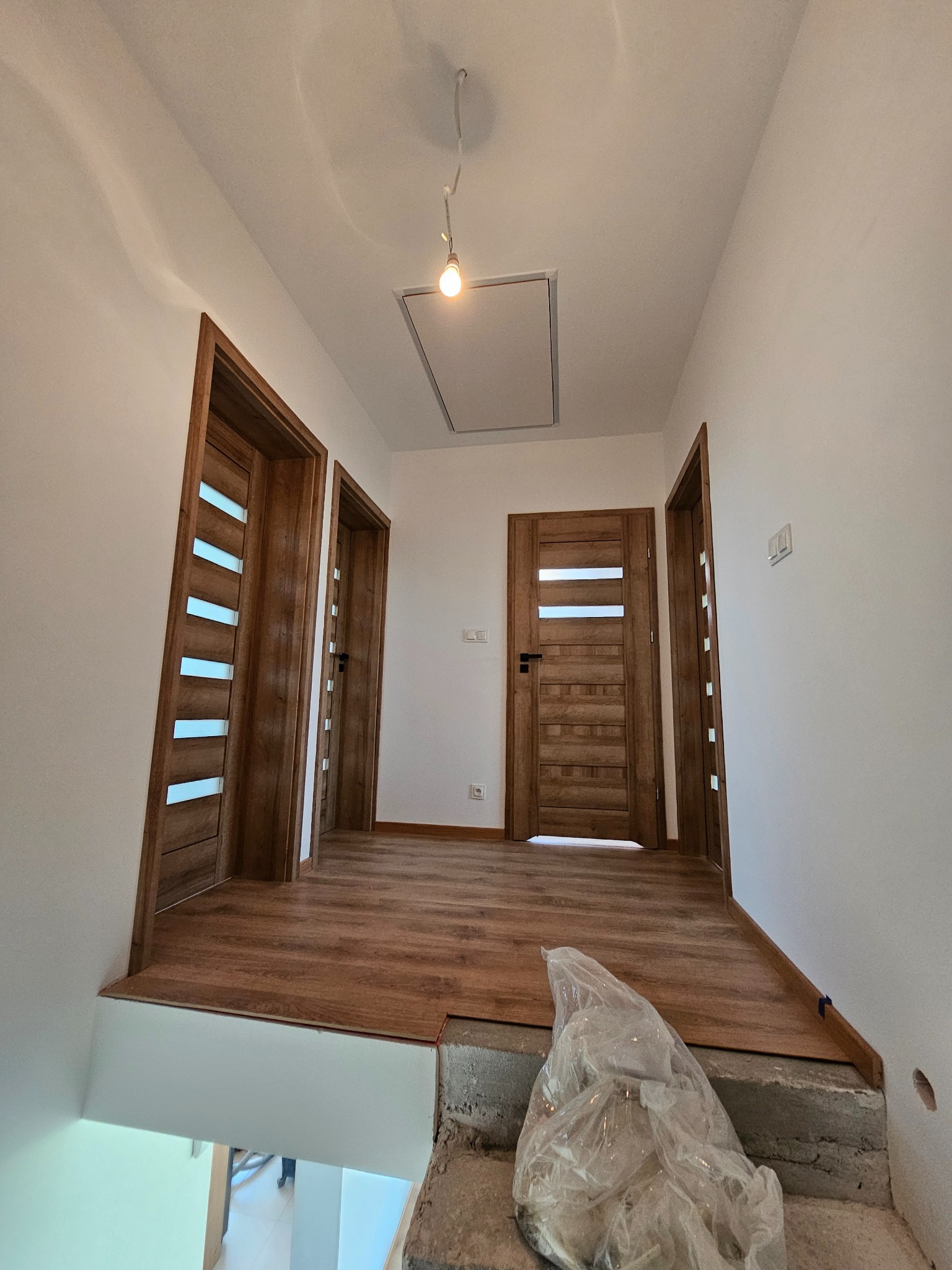 Wnętrze w trakcie wykańczania: drewnopodobna podłoga, trzy drewniane drzwi z przeszkleniami, białe ściany. Widoczny fragment schodów w stanie surowym.