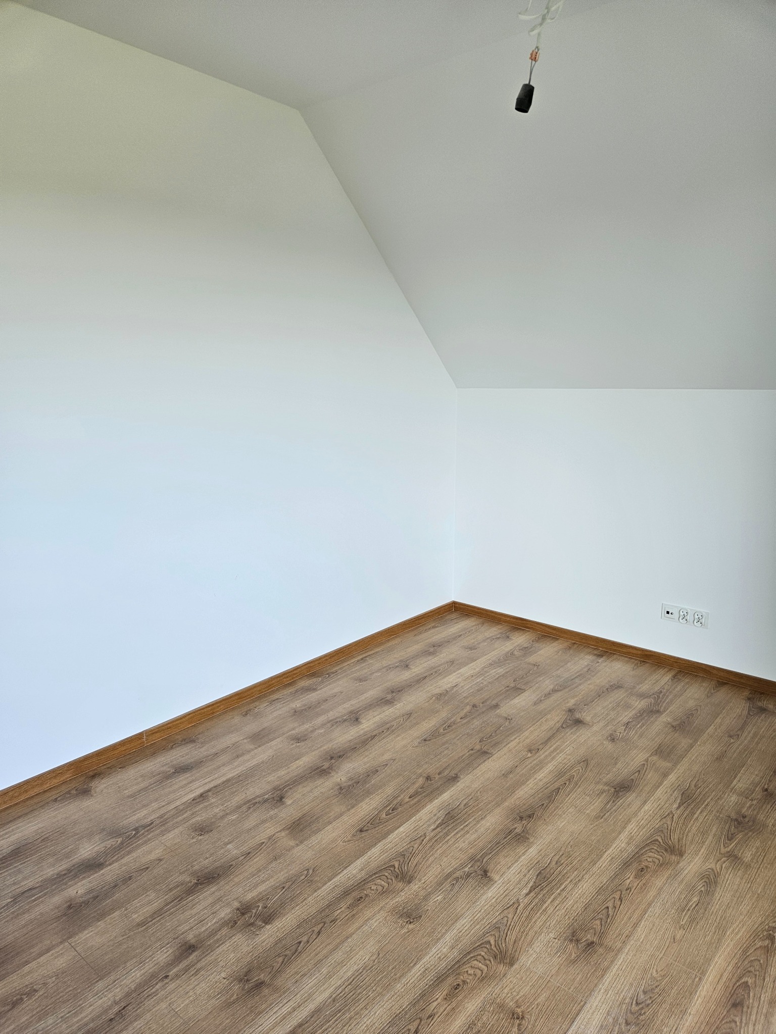 Jasne panele podłogowe ułożone w pokoju na poddaszu z białymi ścianami i widocznymi gniazdkami elektrycznymi. W tle widoczny kabel od lampy.