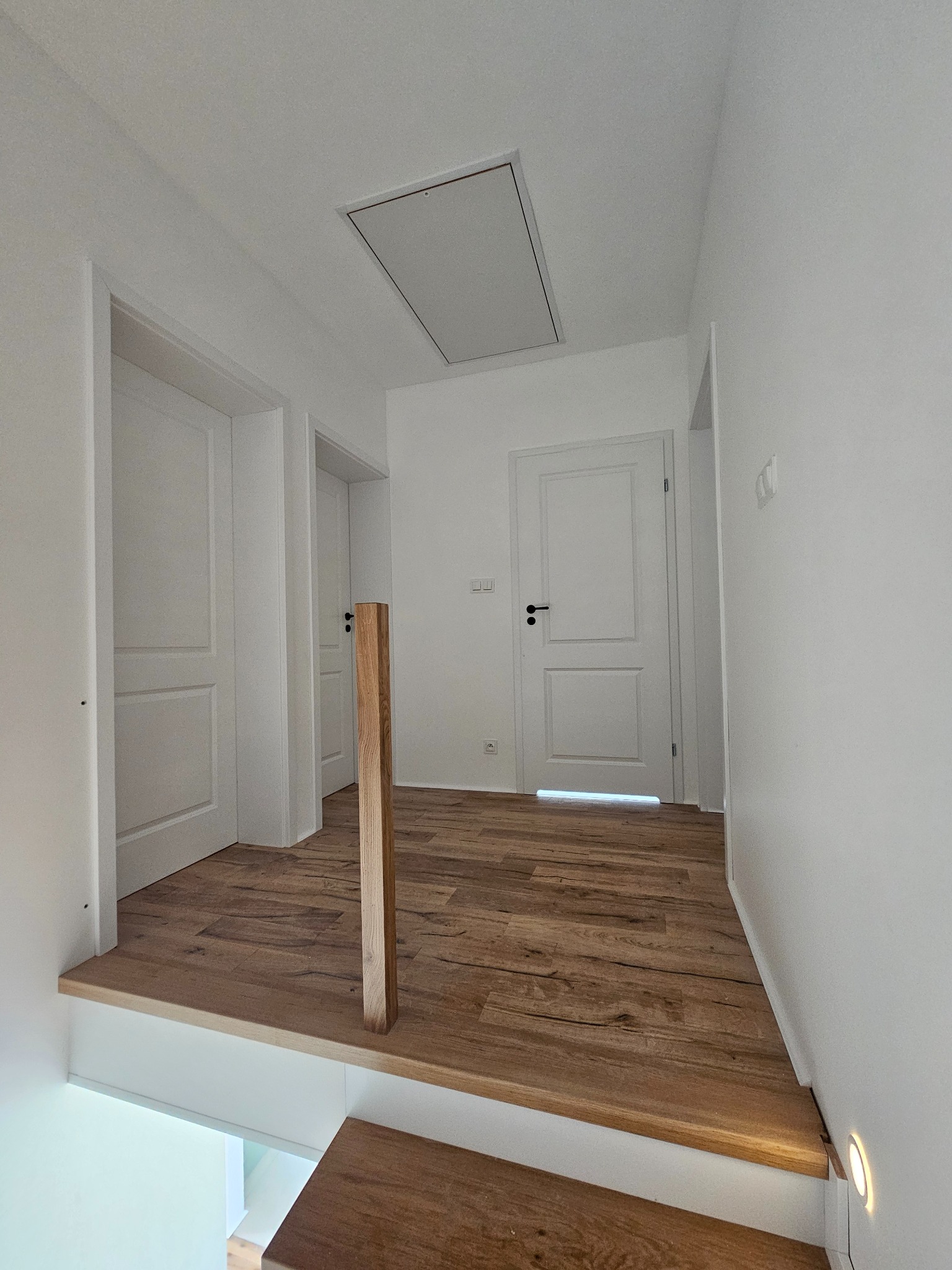 Jasne wnętrze po remoncie: drewniana podłoga, białe ściany i drzwi. Widoczne schody z podświetleniem LED i drewniana barierka. Minimalistyczny design.