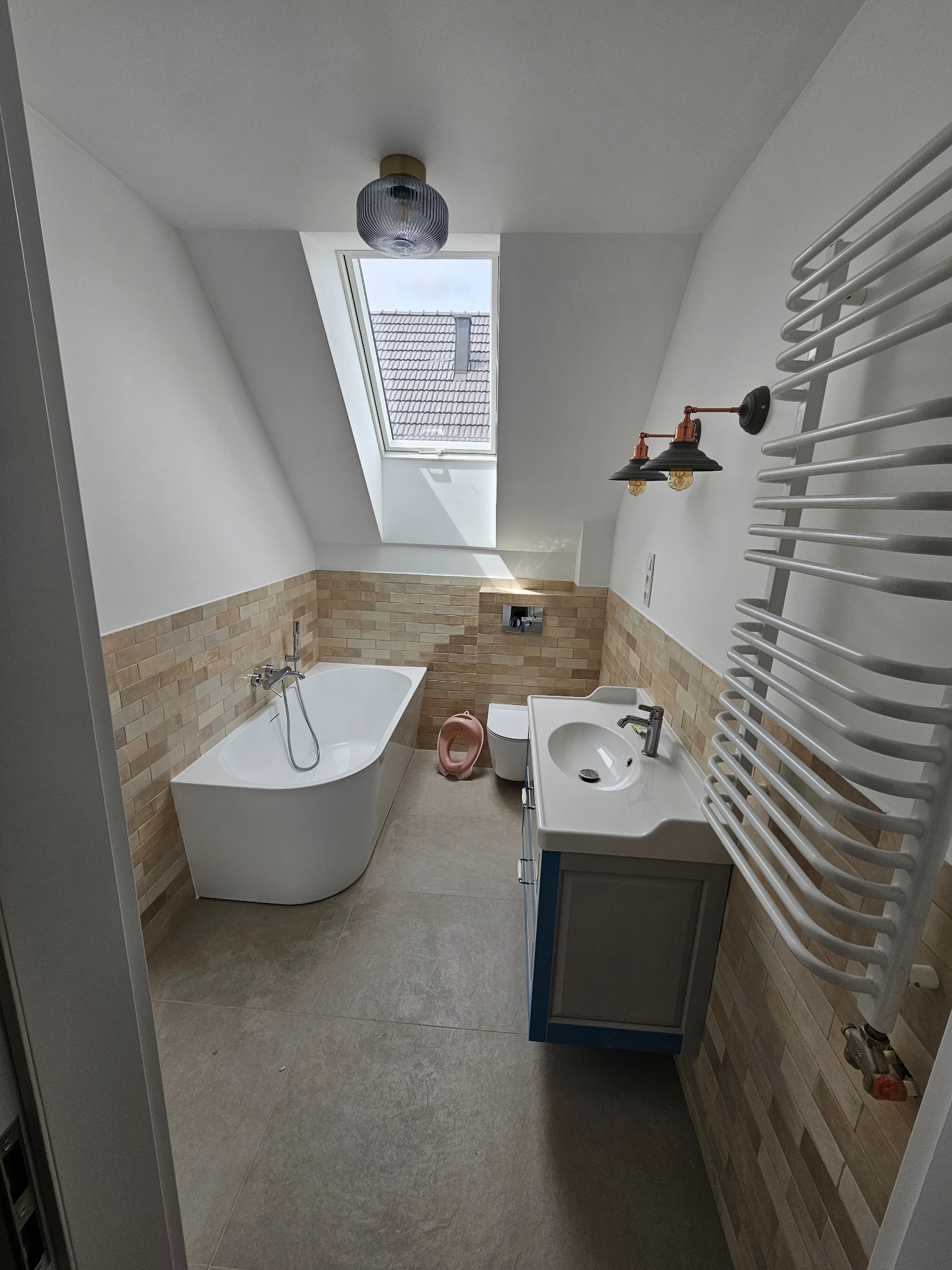Jasna łazienka na poddaszu z beżowymi płytkami, białą wanną i umywalką z szafką. Widoczne okno dachowe i grzejnik drabinkowy. Styl nowoczesny z elementami retro.