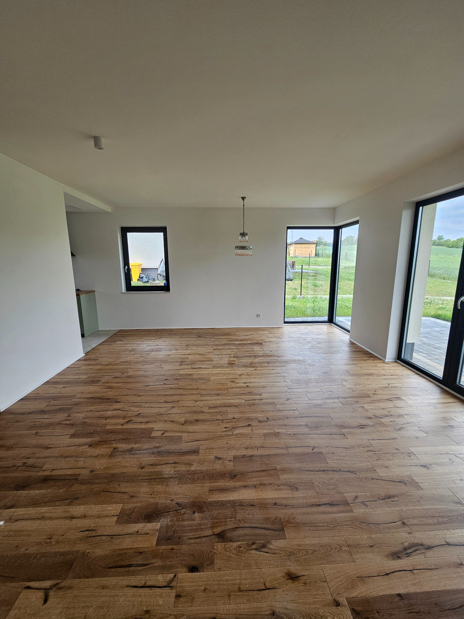 Jasne wnętrze z drewnianą podłogą, dużymi oknami i minimalistycznym oświetleniem. Widok na zewnątrz z zielenią. Nowoczesny design i przestronność.