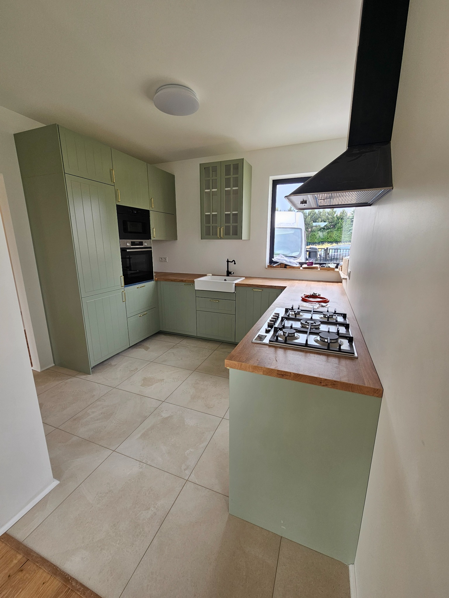 Jasna kuchnia w stylu rustykalnym z zielonymi szafkami, drewnianym blatem i białym zlewem farmerskim. Czarny okap nad płytą gazową. Beżowe płytki podłogowe.