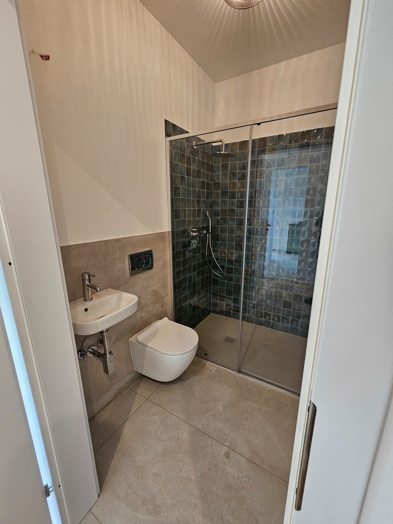 Nowoczesna łazienka z kabiną prysznicową z ciemnymi płytkami, podwieszaną toaletą i umywalką. Minimalistyczny design, jasne płytki podłogowe, widok zza otwartych drzwi.