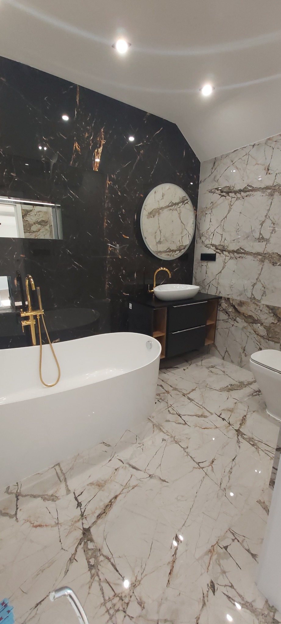 Elegancka łazienka z czarnymi i białymi marmurowymi płytkami, wolnostojącą wanną i okrągłym lustrem nad umywalką. Złote akcenty armatury dodają luksusowego charakteru.