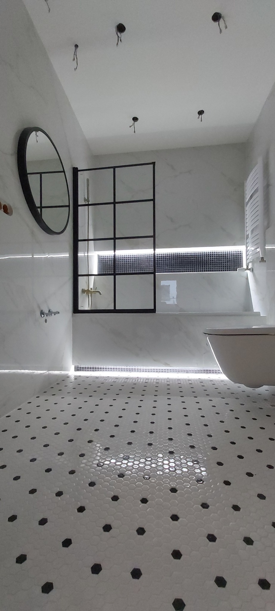 Nowoczesna łazienka z heksagonalną podłogą, czarną ramą kabiny prysznicowej i okrągłym lustrem. Oświetlenie LED podkreśla marmurowe ściany i armaturę.