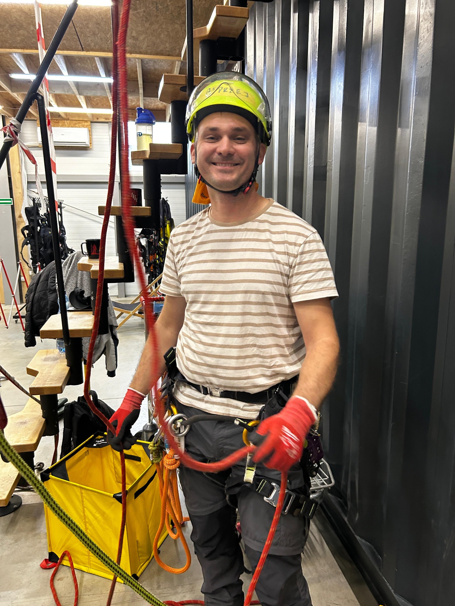 Uśmiechnięty arborysta w kasku i uprzęży, przygotowuje się do pracy z linami. W tle widoczne schody i elementy konstrukcji.