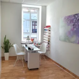 Jasne wnętrze salonu z stanowiskiem do manicure: białe meble, krzesła, lampa, półki z lakierami. Okno z widokiem na budynek. Minimalistyczny wystrój, drewniana podłoga.