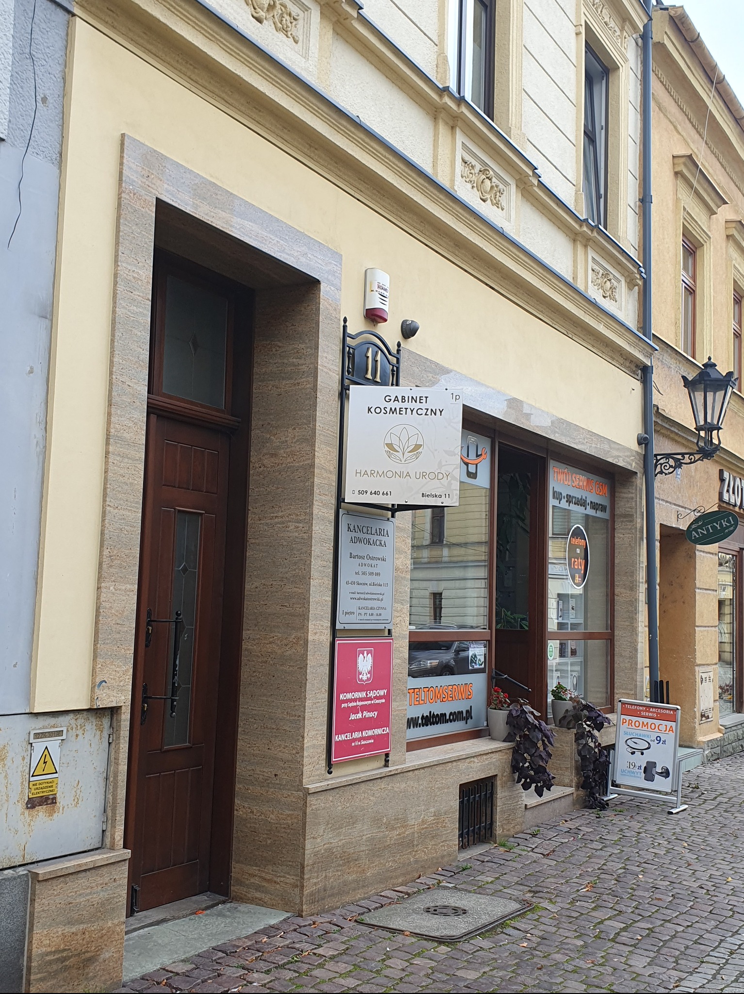 Wejście do gabinetu kosmetycznego 'Harmonia Urody' w kamienicy, z szyldem i reklamą usług GSM. Widok z brukowanej ulicy, w tle antykwariat.