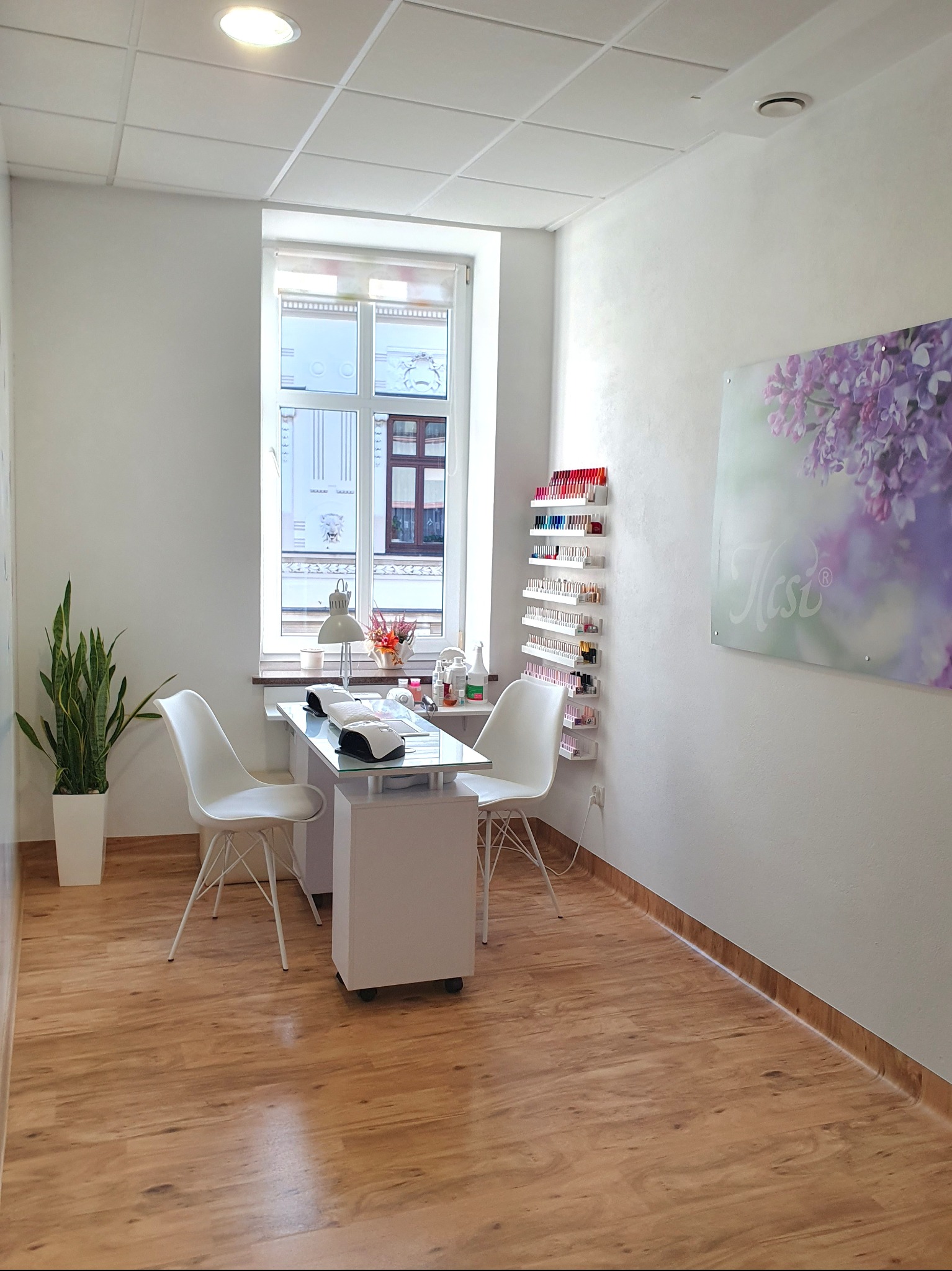 Jasne wnętrze salonu z stanowiskiem do manicure: białe meble, krzesła, lampa, półki z lakierami. Okno z widokiem na budynek. Minimalistyczny wystrój, drewniana podłoga.