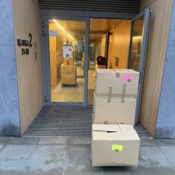 MalardPrzeprowadzki - Klatka schodowa z otwartymi drzwiami, przed którymi leżą kartony z naklejkami, gotowe do transportu. W tle widoczny wózek transportowy z kolejnymi kartonami.