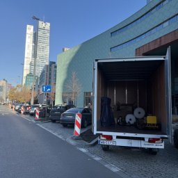 MalardPrzeprowadzki - Otwarty bus transportowy wypełniony meblami i sprzętami na tle miejskiego krajobrazu z wysokimi budynkami. Widoczne zaparkowane samochody i chodnik.