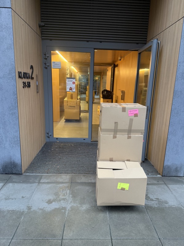 Klatka schodowa z otwartymi drzwiami, przed którymi leżą kartony z naklejkami, gotowe do transportu. W tle widoczny wózek transportowy z kolejnymi kartonami.