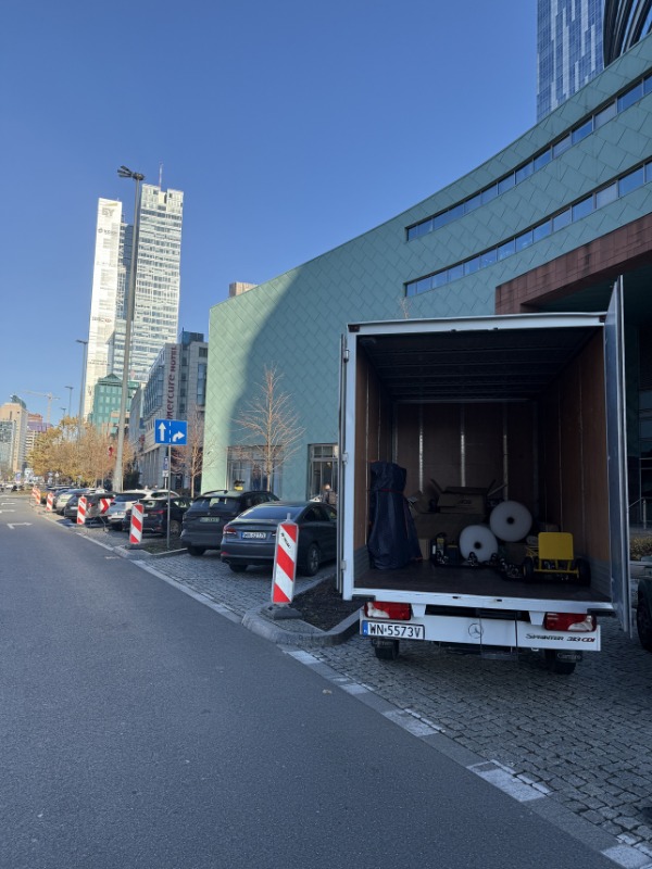 Otwarty bus transportowy wypełniony meblami i sprzętami na tle miejskiego krajobrazu z wysokimi budynkami. Widoczne zaparkowane samochody i chodnik.