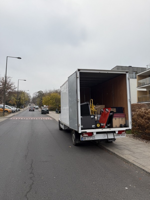 Ciężarówka z otwartymi drzwiami załadowana bagażami i kartonami na ulicy miejskiej, sugerująca przeprowadzkę. Widoczny numer rejestracyjny pojazdu.