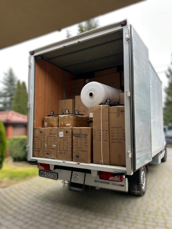 Ciężarówka wypełniona kartonami i folią bąbelkową, gotowa do transportu. Widoczny numer rejestracyjny i logo Sprinter. Detal: koła z rolkami pod kartonami.