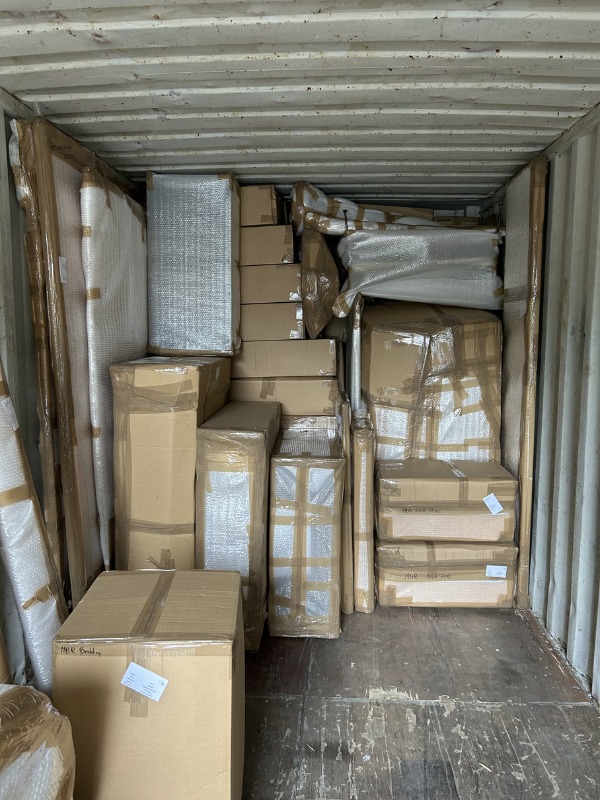 Wnętrze kontenera wypełnione po brzegi kartonami i meblami zabezpieczonymi folią bąbelkową i taśmą, gotowymi do transportu. Widoczne etykiety adresowe na paczkach.
