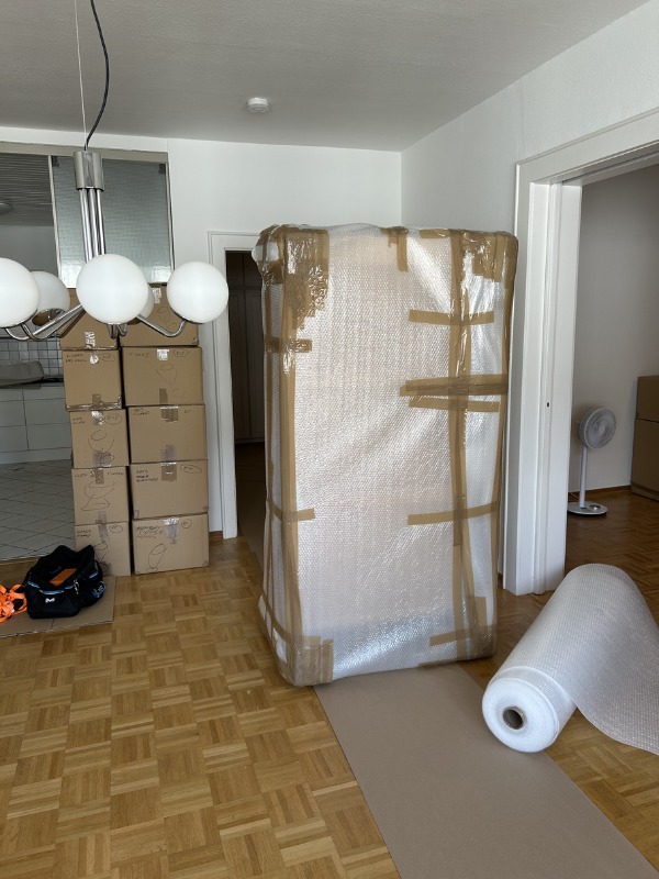 Zabezpieczony mebel folią bąbelkową i taśmą, gotowy do transportu. W tle kartony i lampa sufitowa. Podłoga wyłożona parkietem.