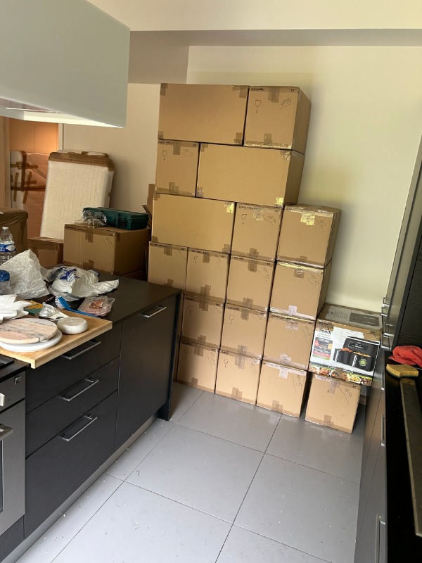 Stos kartonowych pudeł z zawartością, przygotowanych do transportu, w kuchni z szarymi płytkami podłogowymi i czarnymi szafkami. Częściowo spakowane przedmioty na blacie.