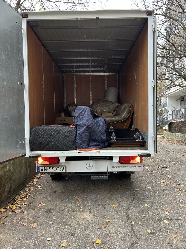 Wnętrze furgonetki Mercedes Sprinter 313 CDI, załadowanej meblami i sprzętami zabezpieczonymi kocami, gotowej do transportu. Widok z tyłu pojazdu na ulicy.