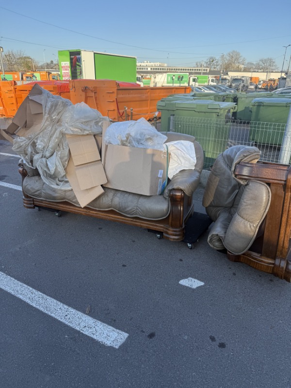 Stary, skórzany fotel z kartonami i folią na parkingu, przygotowany do wywozu. W tle widoczne kontenery i pojazdy komunalne.