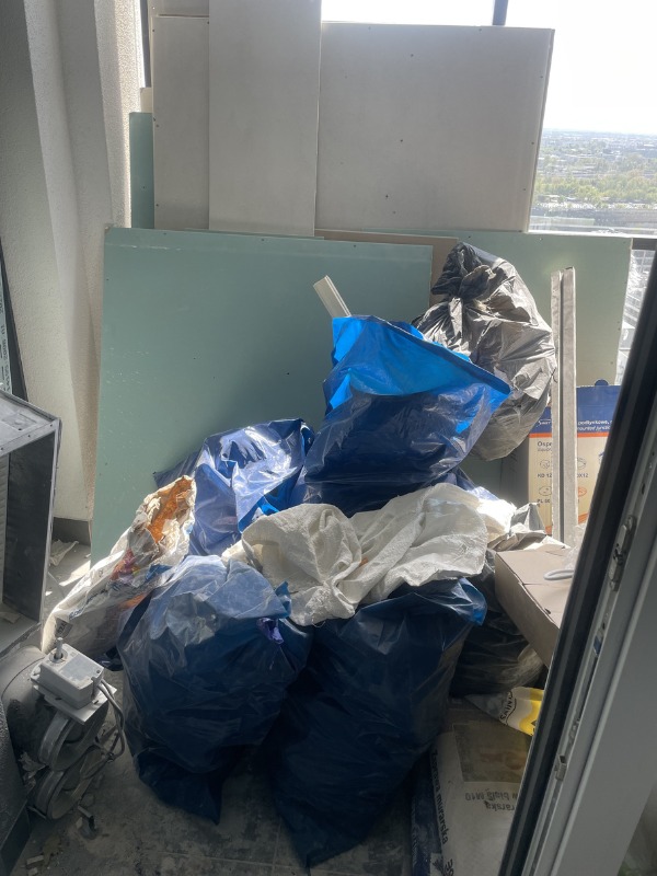 Stos worków z odpadami budowlanymi i płyty gipsowe w pomieszczeniu z widokiem na miasto. Niebieskie i białe worki, gruz, remont, okno.