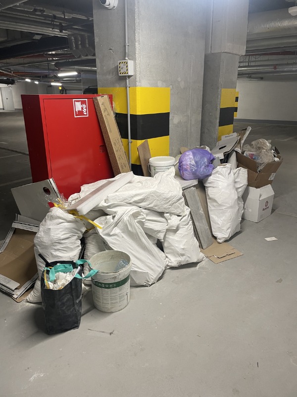 Sterta worków z odpadami budowlanymi i gruzem w garażu podziemnym, obok czerwonej szafki hydrantowej i betonowego filaru z żółto-czarnymi pasami ostrzegawczymi.