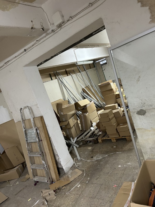 Stare pomieszczenie z kartonami, paletami i drabiną. Widoczne ślady użytkowania i zaniedbania. Przygotowanie do opróżniania lub transportu.