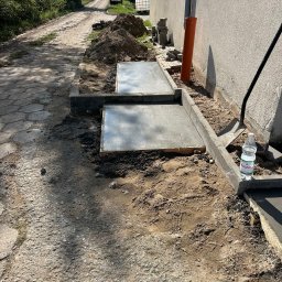 BUD-KOP Adam Świerczewski - Wylane fundamenty pod budynek z obrzeżami, widok z boku. Na pierwszym planie droga gruntowa, w tle budynek i drzewa. Prace ziemne w toku, butelka wody obok.