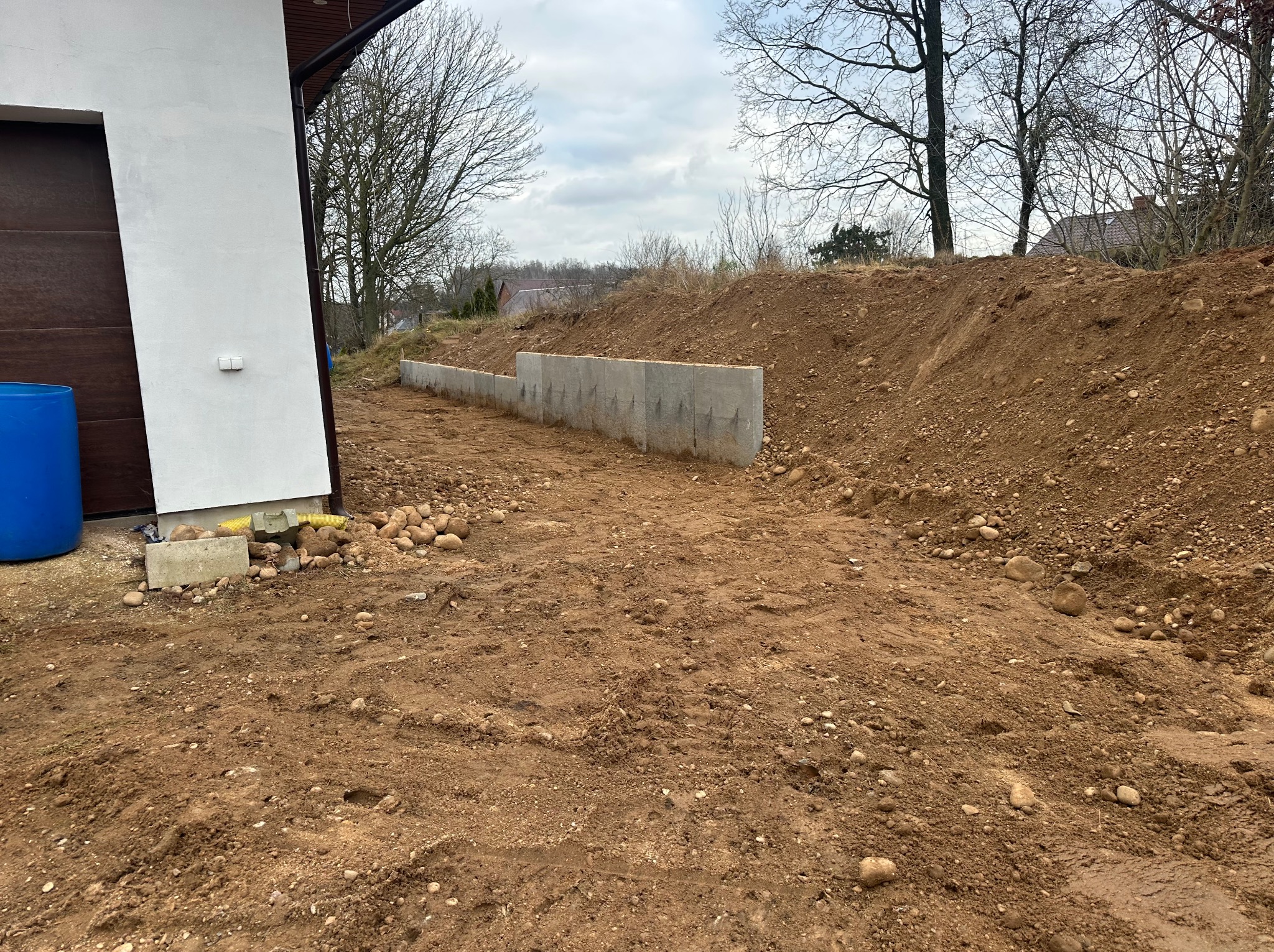 Wzniesiony mur oporowy z betonowych bloczków na nierównym terenie z ziemią i kamieniami, obok fragment budynku z brązową bramą garażową i niebieskim zbiornikiem.