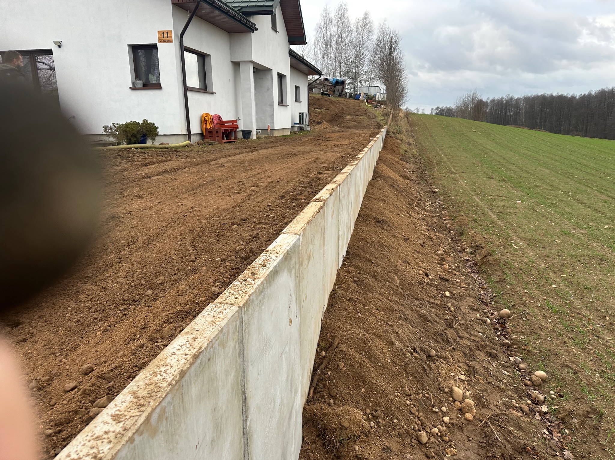 Betonowy murek oporowy na nierównym terenie, zabezpieczający skarpę przy białym domu z numerem 11. W tle zielone pole i drzewa pod pochmurnym niebem.