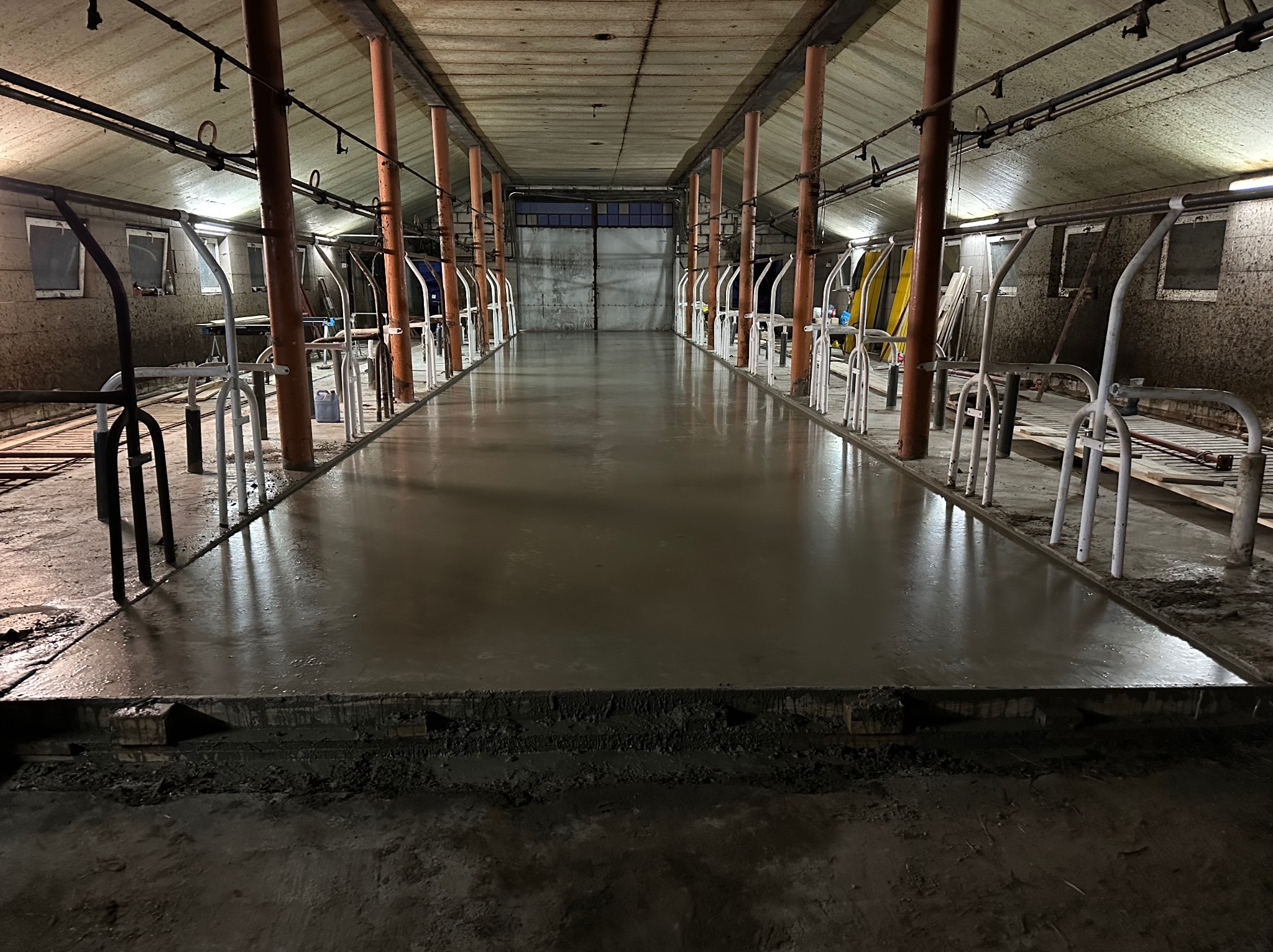 Świeżo wylana, gładka posadzka betonowa wewnątrz budynku gospodarczego z rzędami metalowych konstrukcji. Widoczne słupy i oświetlenie sufitowe.