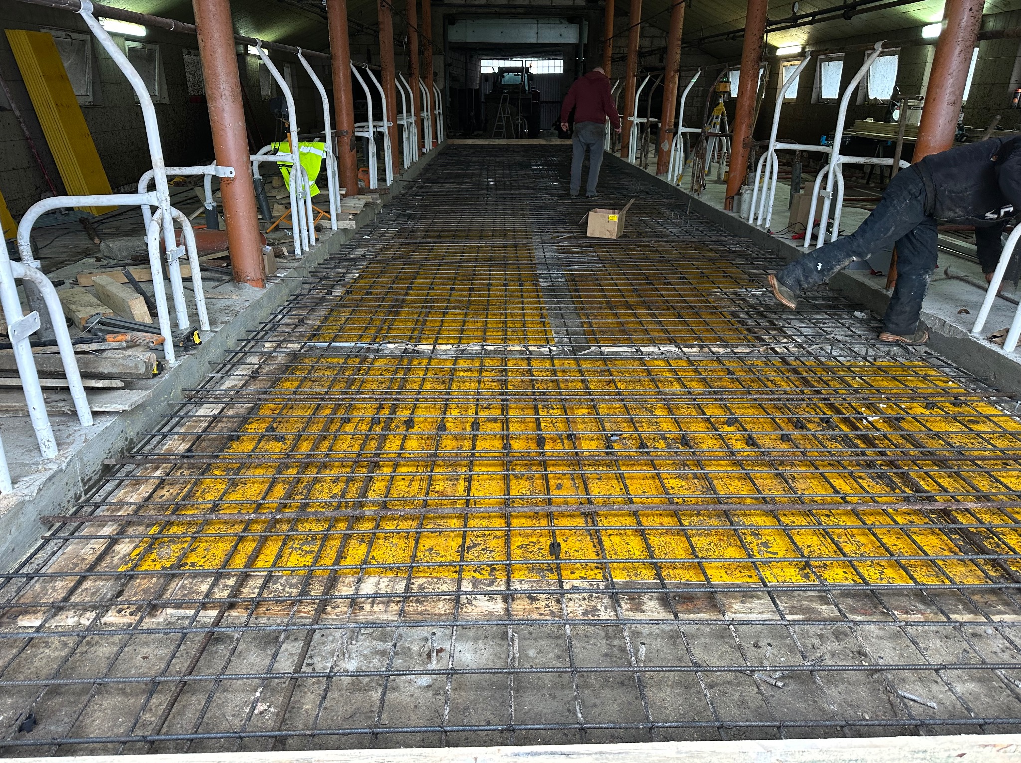 Zbrojenie podłoża wewnątrz budynku gospodarczego, widoczne żółte deski szalunkowe i stalowe pręty tworzące siatkę. W tle osoby i elementy konstrukcyjne.