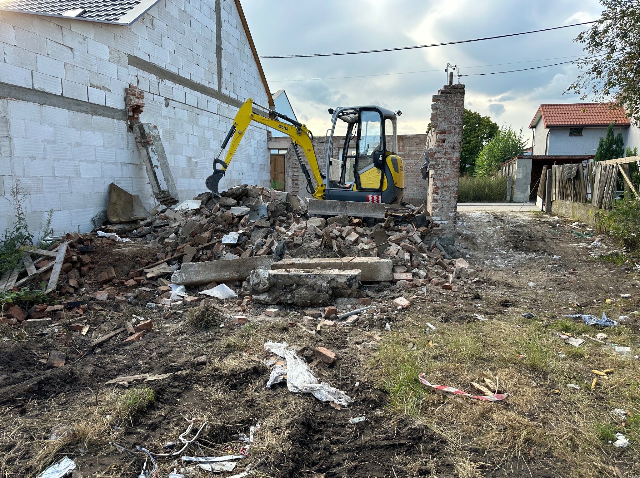 Żółta minikoparka na stercie gruzu po rozbiórce ściany z białego bloczka. Widoczne fragmenty cegieł i resztki budynku w tle. Niebo pochmurne.