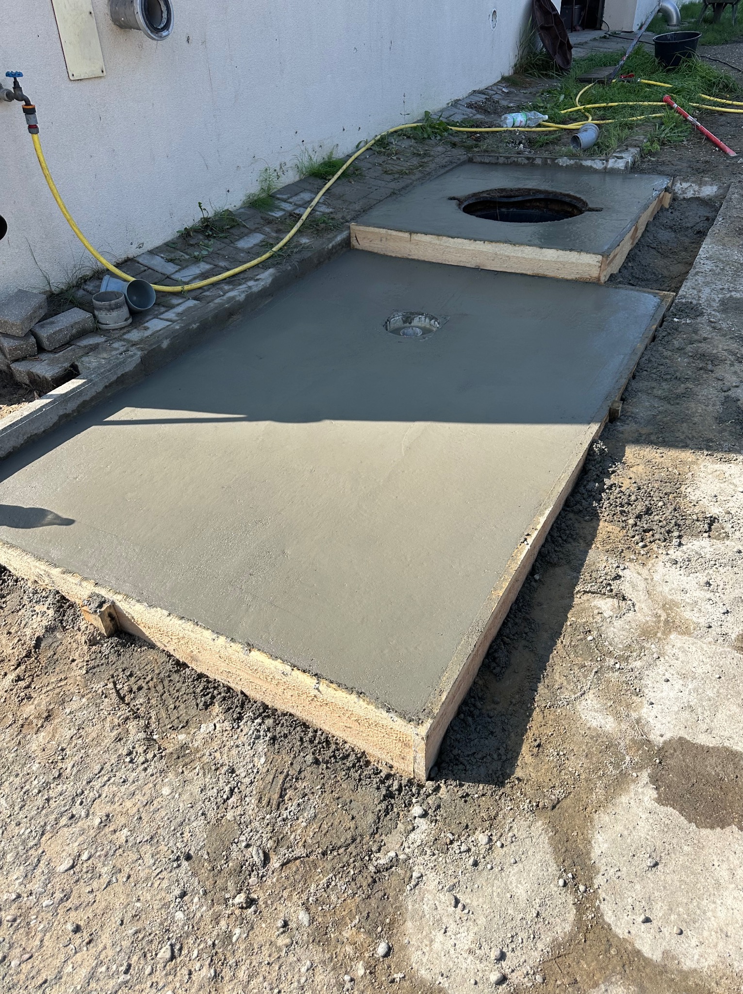 Świeżo wylany beton na szambo z drewnianym szalunkiem, widoczny otwór rewizyjny. Żółty wąż ogrodowy leży obok. Praca w toku, teren budowy.