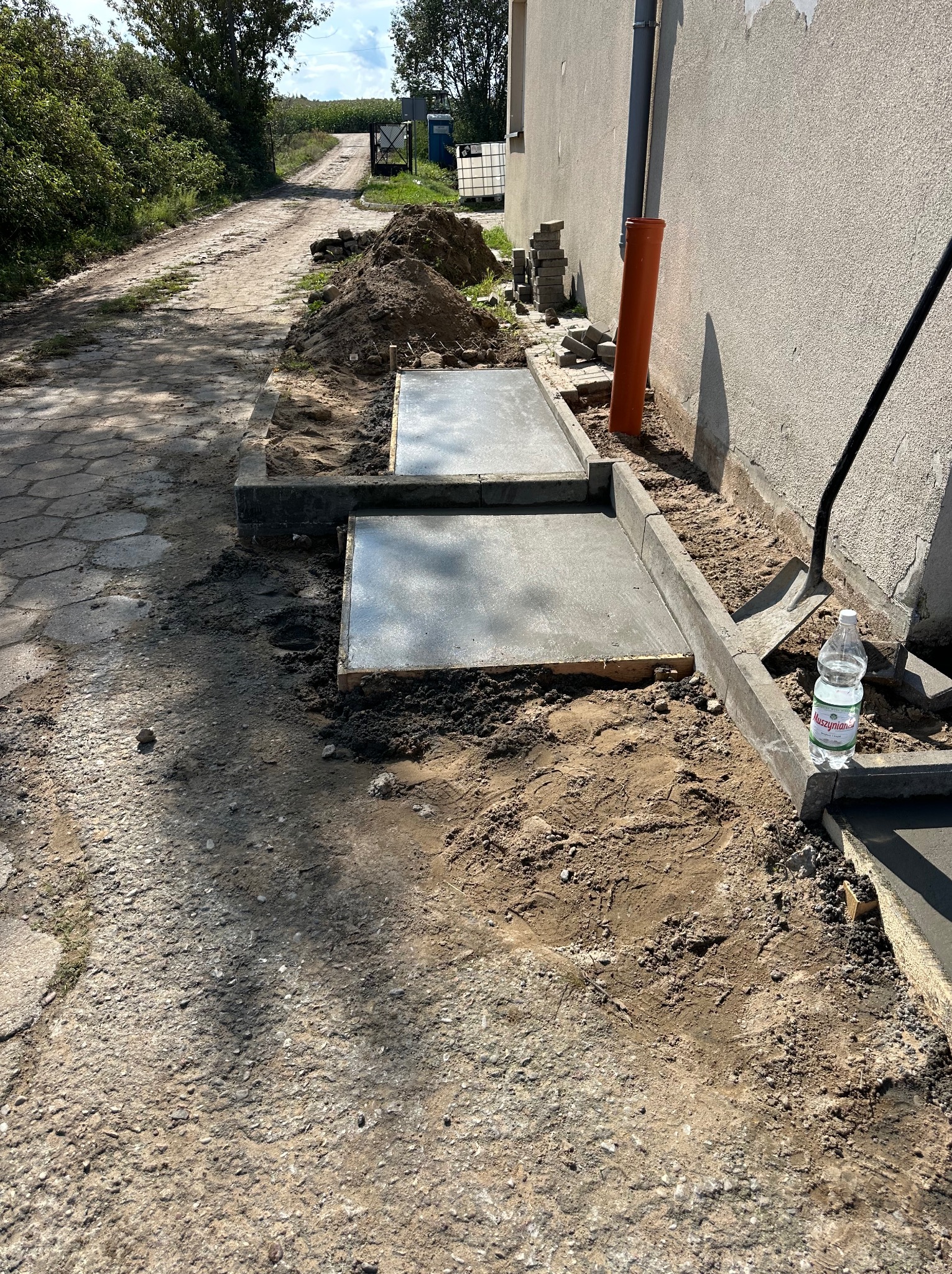 Wylane fundamenty pod budynek z obrzeżami, widok z boku. Na pierwszym planie droga gruntowa, w tle budynek i drzewa. Prace ziemne w toku, butelka wody obok.