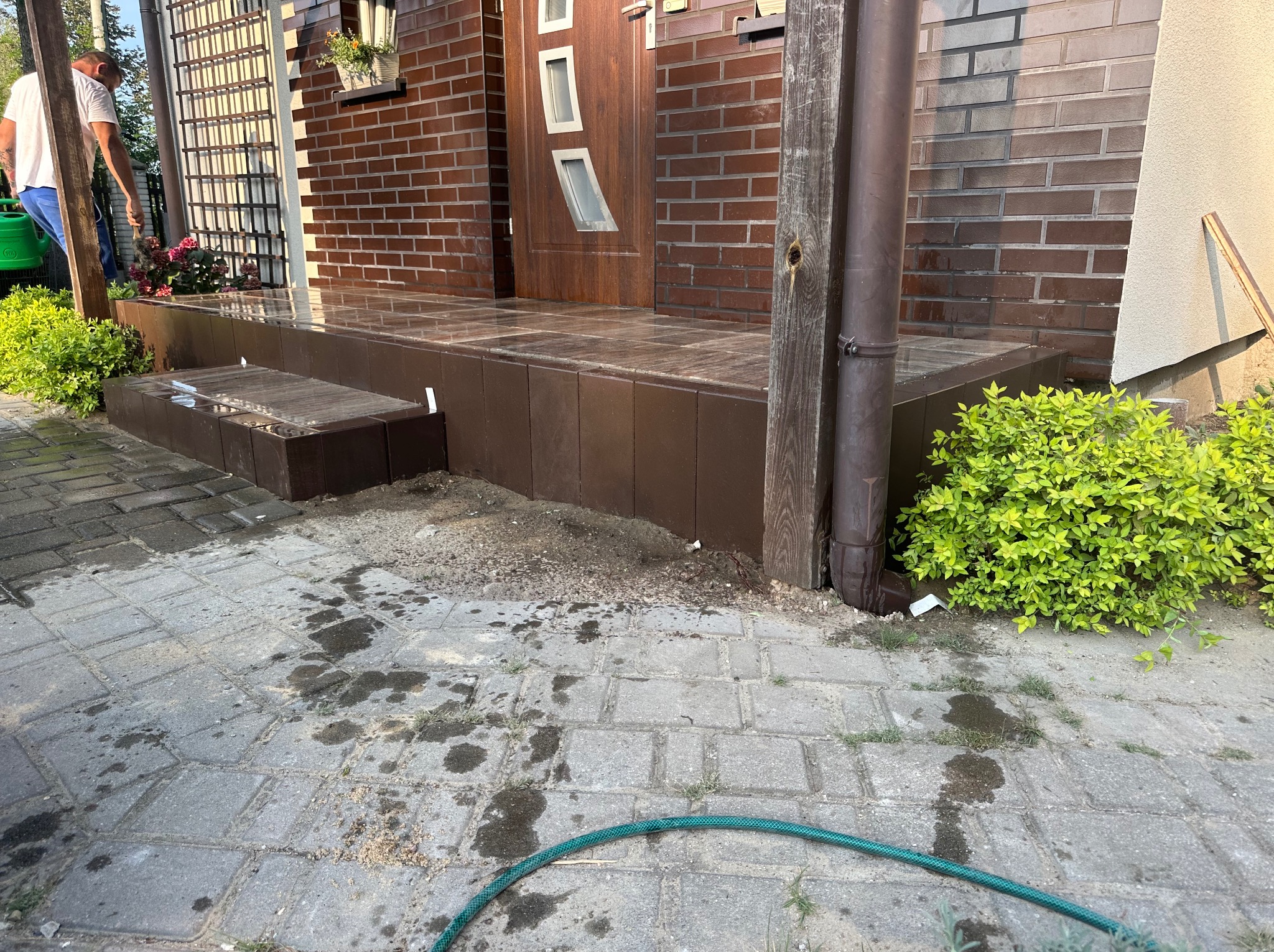 Murowany, brązowy ganek z płytkami przed wejściem do domu w Łomży. Widoczny fragment elewacji z cegły i drewniane drzwi z przeszkleniami.