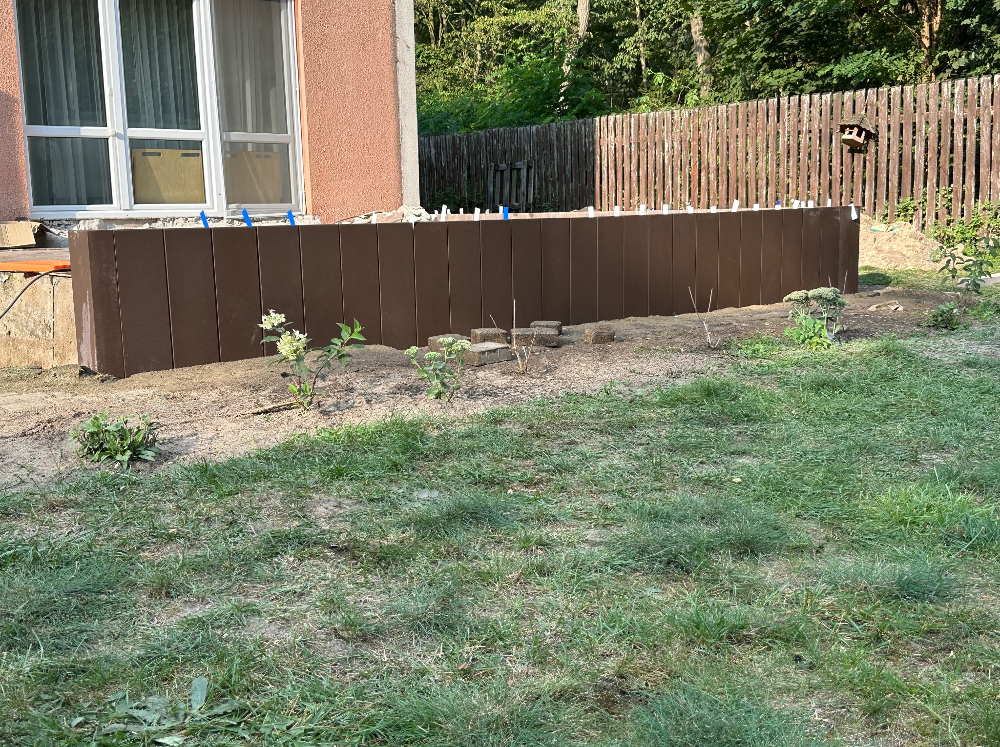 Brązowy murek oporowy z kompozytu, zabezpieczający skarpę przy domu z oknem. W tle drewniany płot i zieleń. Widoczne rośliny i trawa.