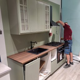 SQUAD S.C. JACEK BARTKOWIAK I KAROL KŁOSEK - Wrocław: Montaż szafek kuchennych z przeszklonymi frontami i drewnianym blatem. Widoczny pracownik instalujący okap. Elementy szuflad czekają na montaż.
